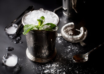 Mint julep whiskey cocktail in misted frozen metal glass and bar accessories shaker jigger spoon ice sugar on black background. Restaurant menu, alcohol recipe decor, specialized website blog design