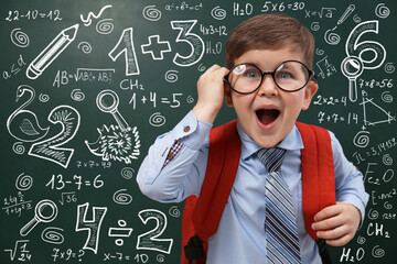 Funny little child wearing glasses near chalkboard with mathematical equations. First time at school