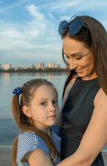 Fototapeta na wymiar Mother and daughter stand close to each other at the lake shore. The mother smiles at her daughter, and the girl looks at the camera. Both are beautiful, of European appearance. High quality photo