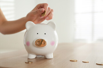 Woman putting money into piggy bank at wooden table indoors, closeup. Space for text