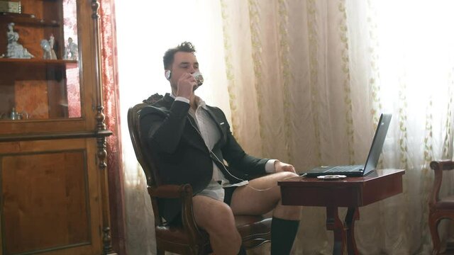 businessman working from his home through a laptop half-dressed in a suit and underpants drinks coffee and waves his hand at the computer