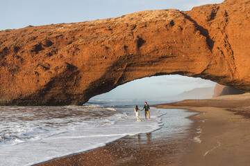 Couple in love walking under huge arch on Legzira ocean beach at sunset. Famous and popular...