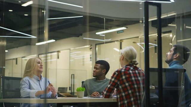 Young man with disposable coffee cup entering office with glass walls and presenting ideas on papers to group of diverse colleagues sitting at table, listening to him and brainstorming in meeting