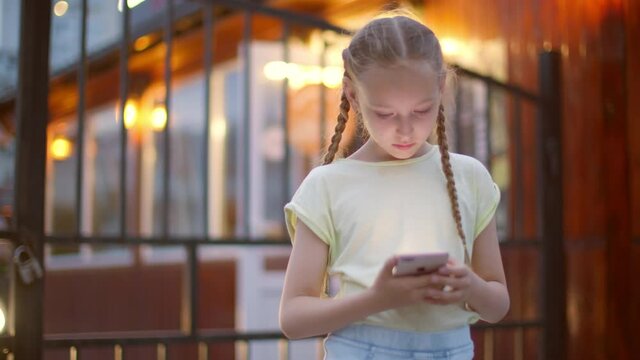Teenager girl using mobile phone at evening outdoor. Young girl with two braids browsing smartphone on evening city street