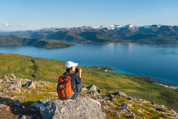 Young Asian woman traveller with backpack sitting on rock taking picture of Senja island in summer season, Norway, Scandinavia