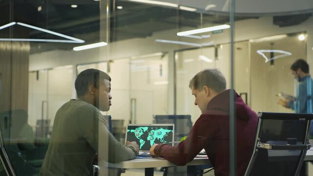 Back view of Black man and his Caucasian colleague sitting at desk with open laptop with world map on screen and discussing marketing strategy behind glass in office, people walking in background