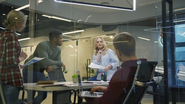Panning shot of diverse group of five business people discussing ideas in office behind glass wall. Middle aged woman explaining her opinion to colleagues