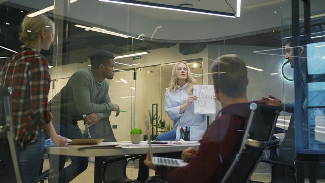 Zoom in shot of diverse group of five business people brainstorming in office behind glass wall. Middle aged woman presenting her ideas to colleagues