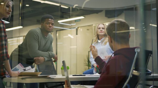 Zoom in shot of young man using laptop computer and sharing ideas with diverse business team in meeting behind glass wall