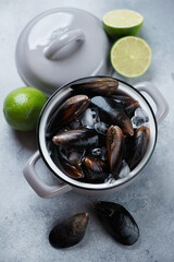 Fresh uncooked iced mussels with limes in a grey pan, vertical shot on a light-blue stone background