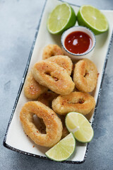 Close-up of fried breaded calamari rings with lime and dipping sauce served on a white plate, vertical shot, selective focus