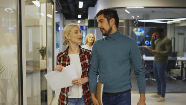 Medium shot of young woman with papers and her male colleague with notepad walking down office corridor and talking cheerfully