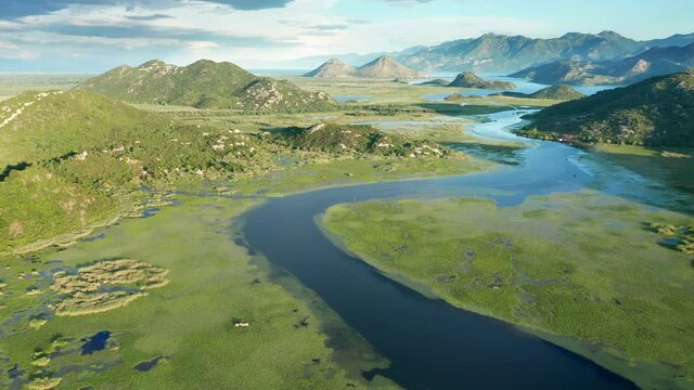 Blue river running through green valley toward distant mountains. Bends and curves of Crnojevica river in Montenegro, meandering through the marsh, on its way to lake Skadar. Aerial footage.