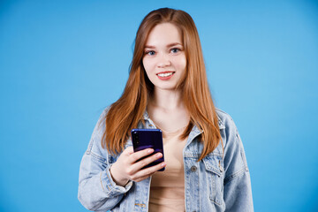 Ginger girl in t-shirt and denim jacket posing isolated on blue wall background. Using mobile phone, typing sms message