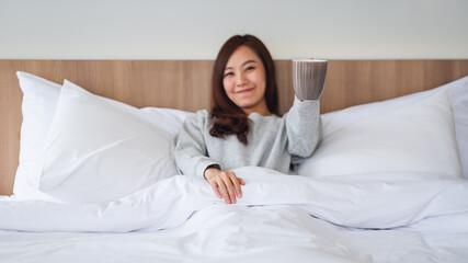 A beautiful asian woman drinking hot coffee while lying on a white cozy bed at home in the morning