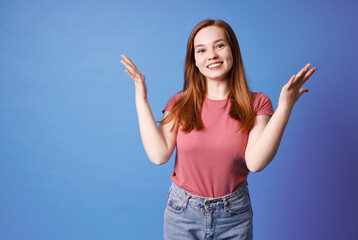 Ginger girl in pink t-shirt and jeans posing isolated on blue. Girl raised her hands, admiring gesture.