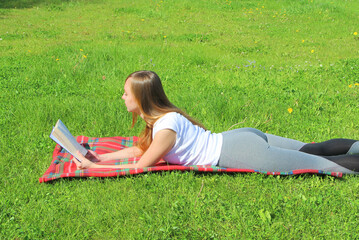 A beautiful young white girl in a white T-shirt and with long hair lies on a red plaid, on green grass, on the lawn and read book