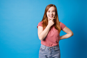 Fototapeta na wymiar Thinking and smiling beautiful young woman in pink t-shirt and jeans posing isolated on blue wall background.