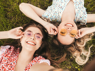 Two young beautiful smiling hipster girls in trendy summer sundress.Sexy carefree women lying on the green grass in sunglasses.Positive models having fun.Top view