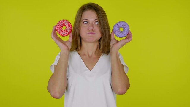 Funny brunette woman in a white T-shirt looks at a delicious blue and red donuts in hands with pouting cheeks, mouth on a yellow background with copy space. Place for text or product. Slow motion