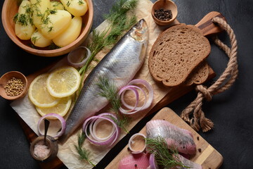 Herring fish  on wooden board  with pepper, herbs, red onion and lemon on black  background. Top view with copy space with potatoes slices and rye bread