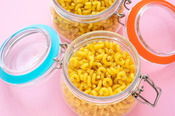 A two glass jars with raw pasta. A pots with yellow macaroni on a pink background.