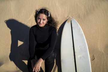 Top view of a smiling female sitting on a sandy hill with a surfboard on the side