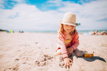 Adorable toddler girl in hat playing on white sand beach at the beach. Summer, vacation.