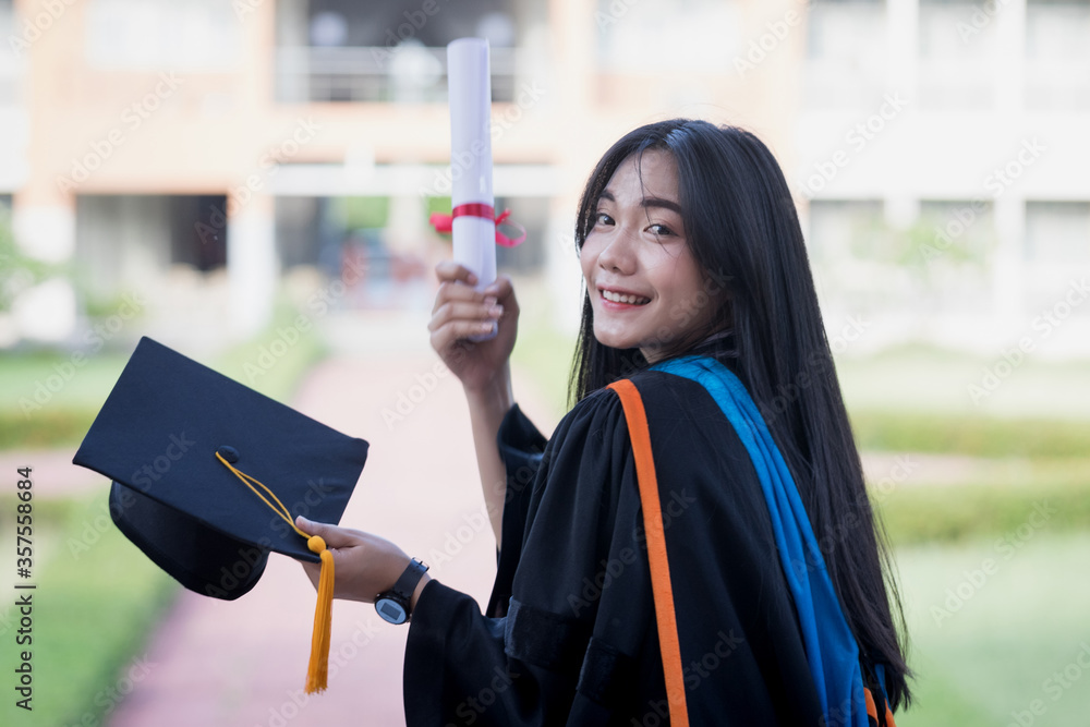 Wall mural Portrait of a happy and excited young Asian female university graduate wears graduation gown and hat celebrates with a degree in the university campus on the commencement day. Education concept.