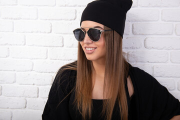 Portrait of young attractive cheerful woman wearing sun glasses and wearing black clothes, studio shot over white bricks background. Happy girl's portrait