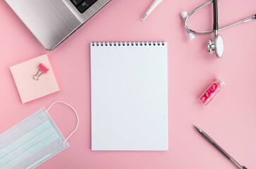 Doctor workplace and medical equipment on pink background. Blank notepad, stethoscope, laptop, protective face mask, thermometer, pills and pen on table. Top view, flat lay, copy space