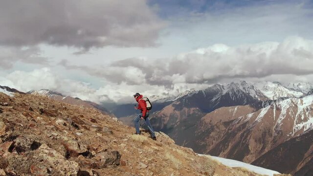 Aerial view of a young thin man with a backpack and trekking sticks climbs uphill on a crest on the border of rocks and rocks with snow against the background of snow-capped peaks and clouds