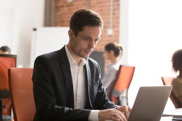 Fototapeta premium Focused employee intern using laptop, working on online project, looking at screen, businessman entrepreneur writing report or business email, analyzing statistics, sitting at office desk