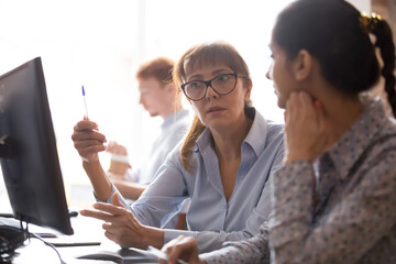 Serious businesswoman mentor teaching new employee, using computer, helping with corporate...