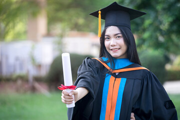 Portrait of a happy and excited young Asian female university graduate wears graduation gown and hat celebrates with a degree in the university campus on the commencement day. Education concept.