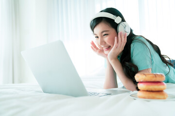 Young girl lying in bed using laptop and wearing headphone to enjoy online content.   