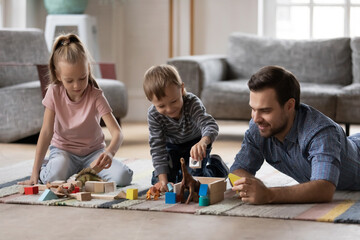 Happy young father with adorable kids playing with toys, colorful wooden blocks, smiling dad with little daughter and son lying on warm floor with underfloor heating at home, enjoying leisure time