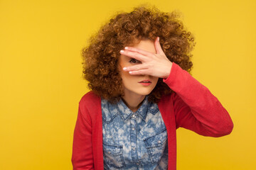 Fototapeta Portrait of curious nosy woman with curly hair spying rumors, looking through fingers with inquisitive expression, shy and scared to watch secret. indoor studio shot isolated on yellow background obraz