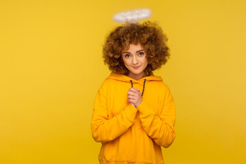Portrait of kind lovely curly-haired hipster woman with saint nimbus holding hands in prayer and...