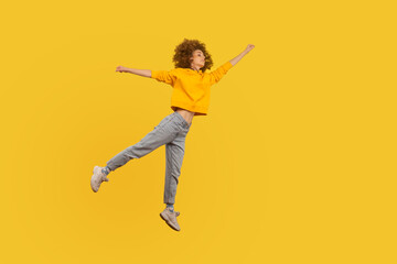 Fototapeta na wymiar Portrait of superhero curly-haired girl in urban style outfit flying with raised hand high in air, feeling superpower and inspiration, achieving goal. indoor studio shot isolated on yellow background