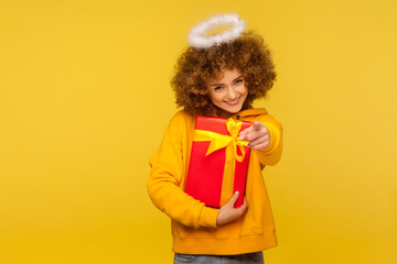 Hey you, happy holidays! Portrait of cheerful curly-haired angelic woman with saint nimbus pointing...