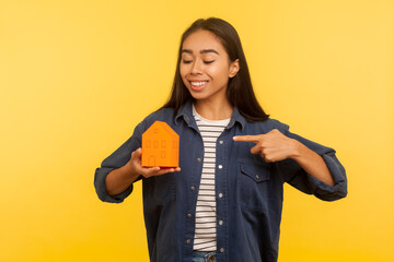 My own home! Portrait of cheerful girl in denim shirt pointing paper house and smiling satisfied, dreaming of real estate purchase, favorable mortgage. indoor studio shot isolated on yellow background