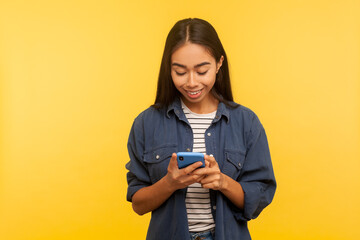 Portrait of happy girl in stylish denim shirt typing message and smiling, dialing number calling on cell phone, using online mobile application, searching web. indoor studio shot, yellow background