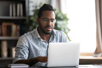 African bearded guy in glasses casual shirt sit at desk at home modern office room using laptop, do...