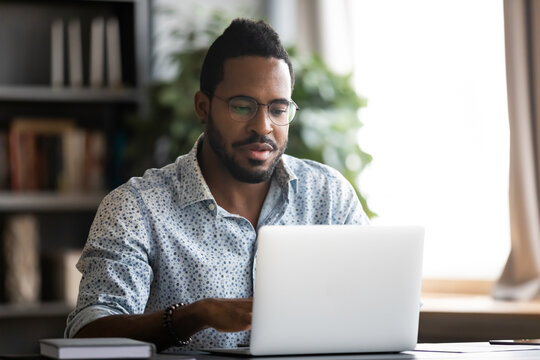 African bearded guy in glasses casual shirt sit at desk at home modern office room using laptop, do remote work, studying distantly passing test online, writing e-mail, websurfing, search info concept