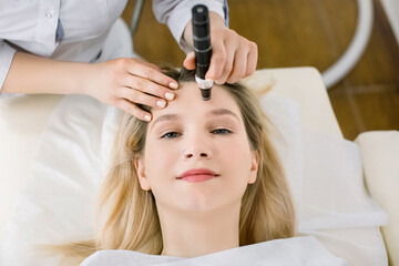 Obraz na płótnie Canvas Dermatology and cosmetology clinic. Cropped top view of face of pretty blond woman during skin diagnostic. Hands of female doctor using a professional dermatoscope while doing the skin examination