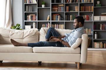 African millennial guy lying barefoot on sofa using wireless computer having fun surfing internet websites enjoy lazy day, chatting via social networks, check e-mail, freelancer do remote work concept