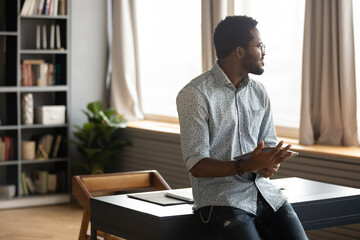 African guy holding tablet distracted from modern wireless device usage looking out the window thinking feels confident and tranquil. Freelancer businessman take break enjoy daydreaming indoor concept