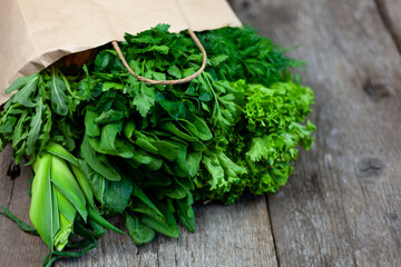 Fresh greens in a paper bag - green onions, dill, parsley, spinach, arugula, sorrel. Home delivery of food and fresh vegetables. Wooden background with place for text. Packaging and cooking.