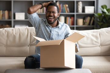 Laughing African guy sitting on couch in living room feels overjoyed opening cardboard box and found confused goods, received unexpected surprise from relatives or friend. Concept of delivery service